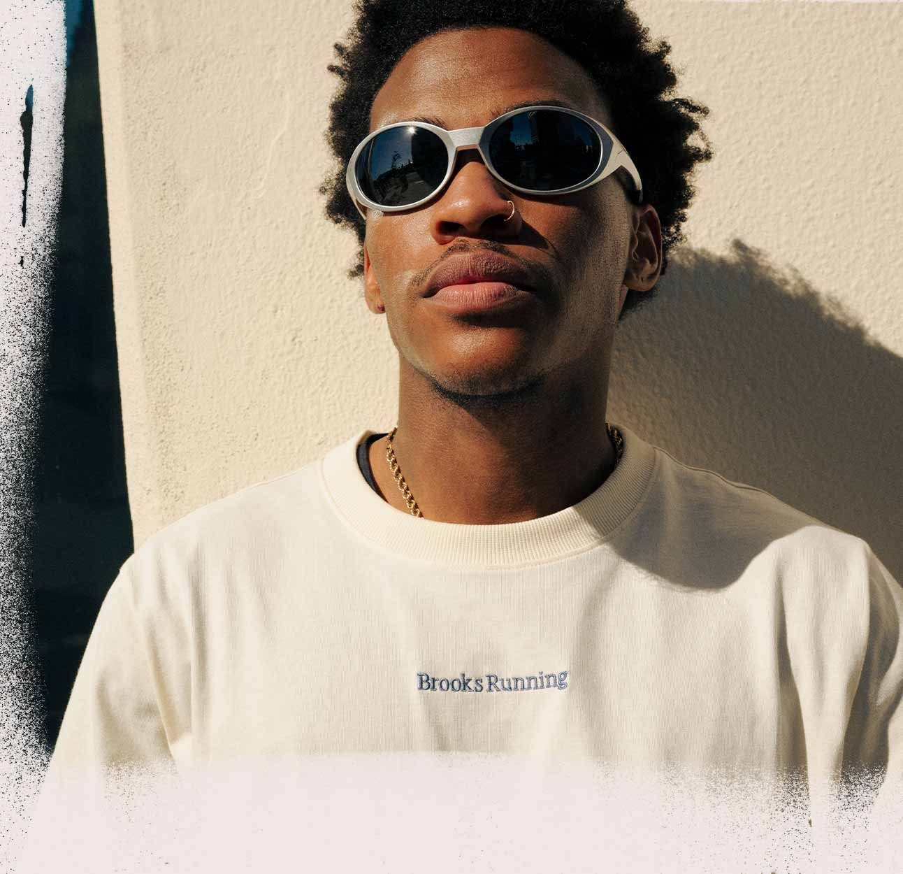 A man wearing a Brooks Running shirt looks ahead, leaning against a white wall